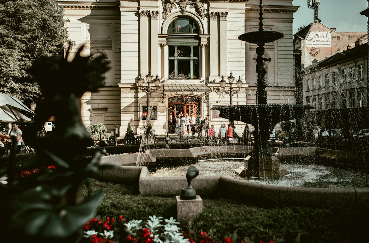 Zdjęcie na zewnątrz. Słoneczny dzień. Na zdjęciu - ozdobna fontanna w kształcie czary i zabytkowa fasada Teatru Polskiego. 