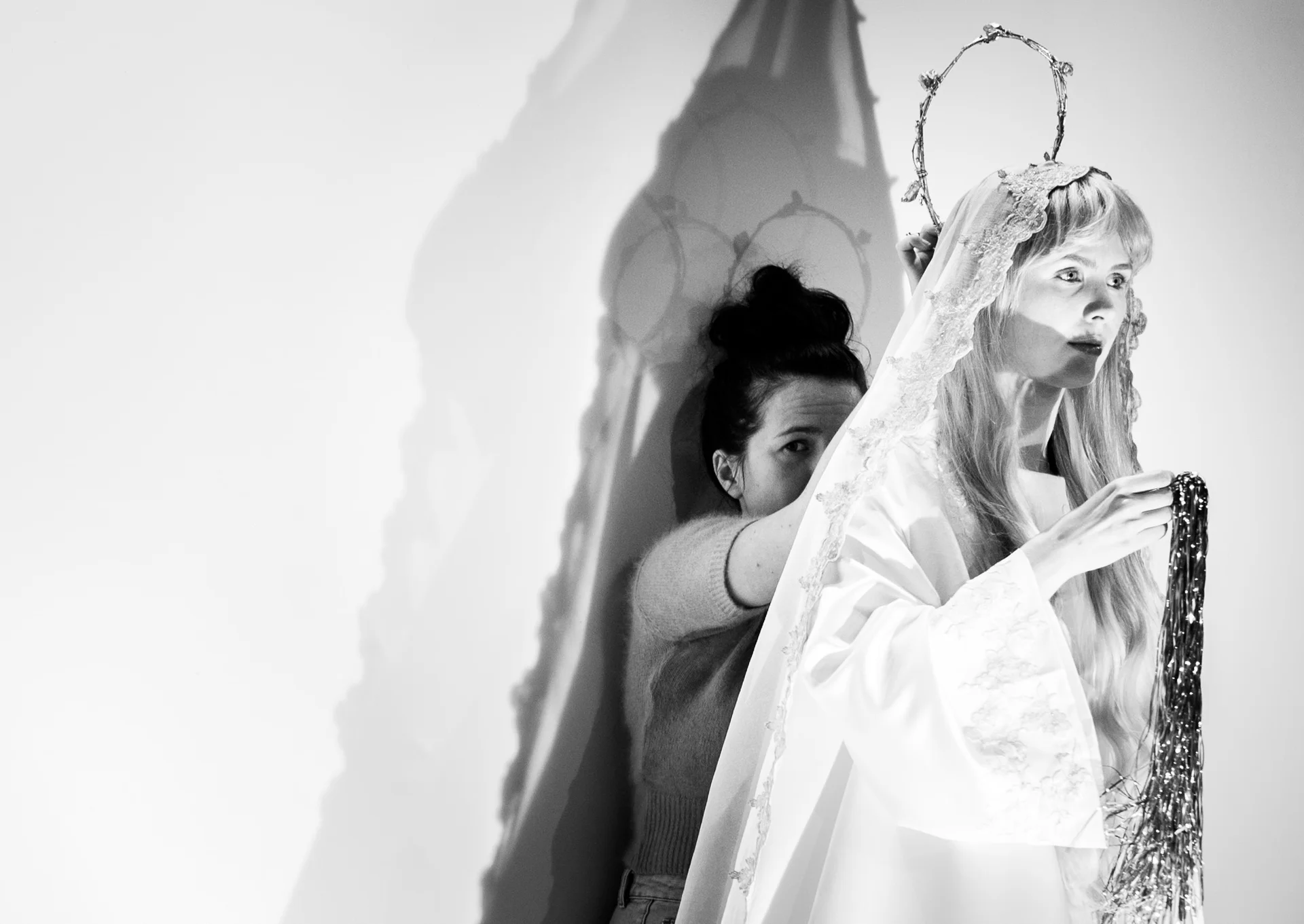 Biało-czarna fotografia. Na białym tle stoi aktorka Oriana Soika stoi ubrana w strój Matki Boskiej. Za nią, w cieniu stoi Iga Sylwestrzak. Trzyma nad głową aktorki drucianą aureolę. 