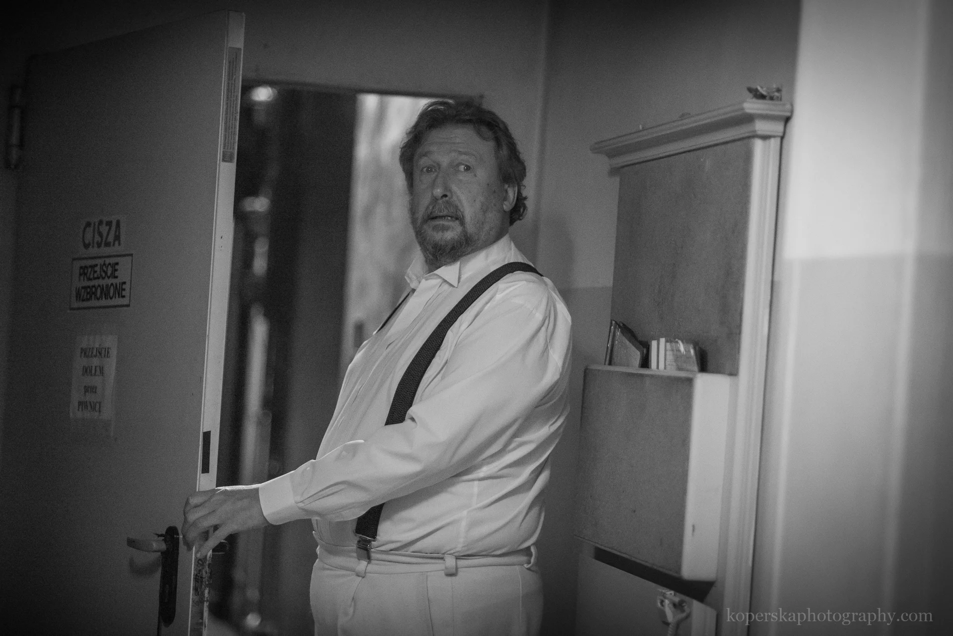 Aktor Kazimierz Czapla stoi w otwartych drzwiach, w wąskim korytarzu. Patrzy wprost w obiektyw. 