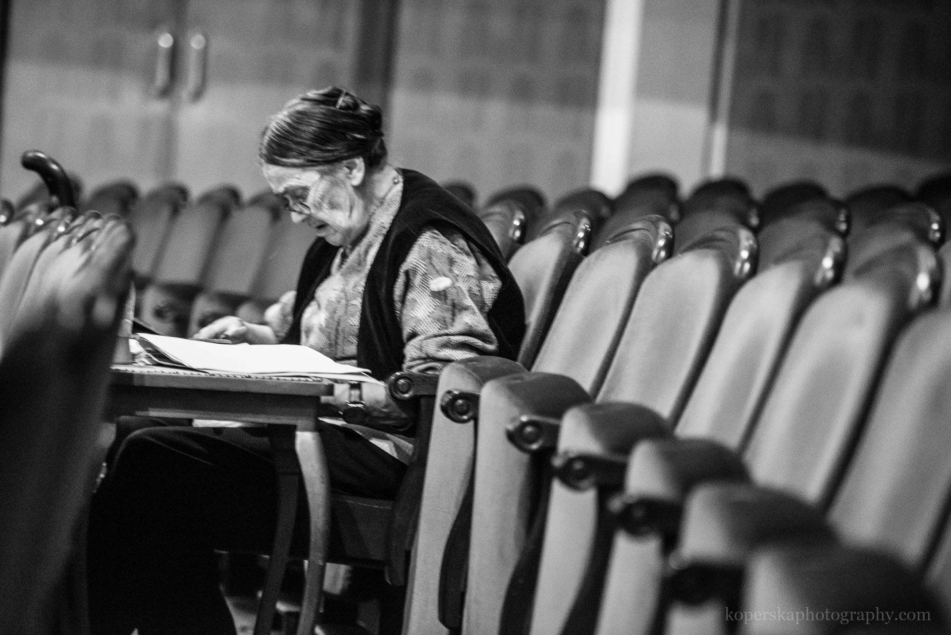 Czarno-białe zdjęcie Małgorzaty Kozłowskiej, która siedzi na widowni teatru. Pochyla się nad otwartym i położonym na kolanach scenariuszem sztuki teatralnej. 