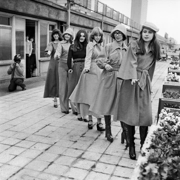 Czarno-białe, archiwalne zdjęcie z lat 70. Sześć młodych kobiet stoi w kolejce, jedna za druga na chodniku przed parterowym budynkiem. 