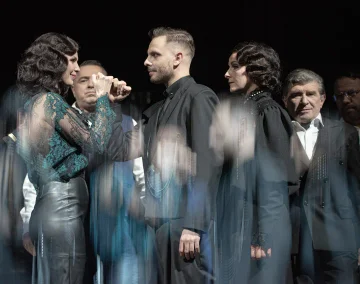 Na scenie stoi grupa osób ubranych na czarno. Na pierwszym planie stoją zwróceni do siebie twarzami Oriana Soika i Michał Czaderna. Trzymają się za ręce i uśmiechają się do siebie. 