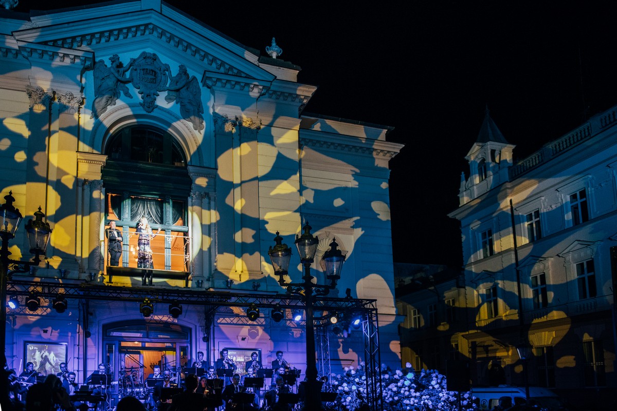 Noc. Fasada Teatru Polskiego oświetlona na kolorowo. Przed wejściem do teatru na krzesłach siedzą muzycy. W oknie, nad  nimi stoi aktorka z mikrofonem. 